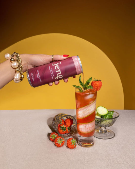 Hand pouring Jeng Rhubarb Cucumber Spritz into a cocktail garnished with strawberry slices and cucumber ribbons.