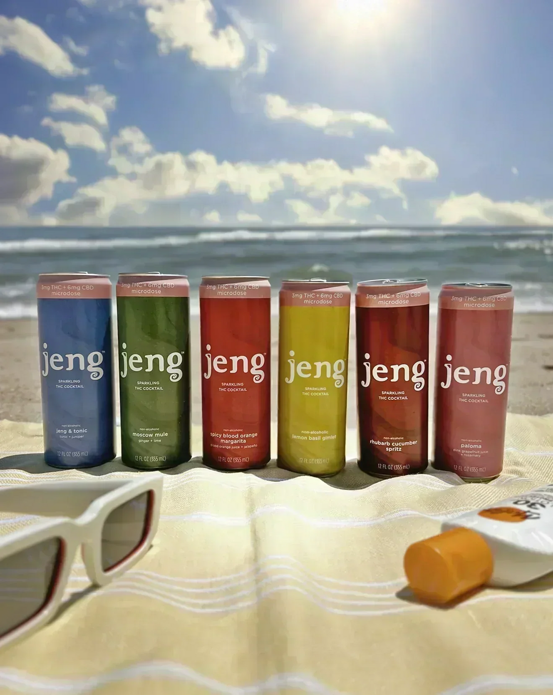 Six Jeng hemp-infused sparkling cocktail cans arranged on a beach towel by the ocean, styled with sunglasses and sunscreen.