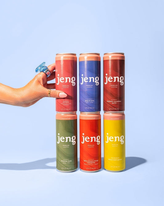 A hand wearing a blue ring touches a stack of six colorful Jeng sparkling cocktail cans against a solid blue background.