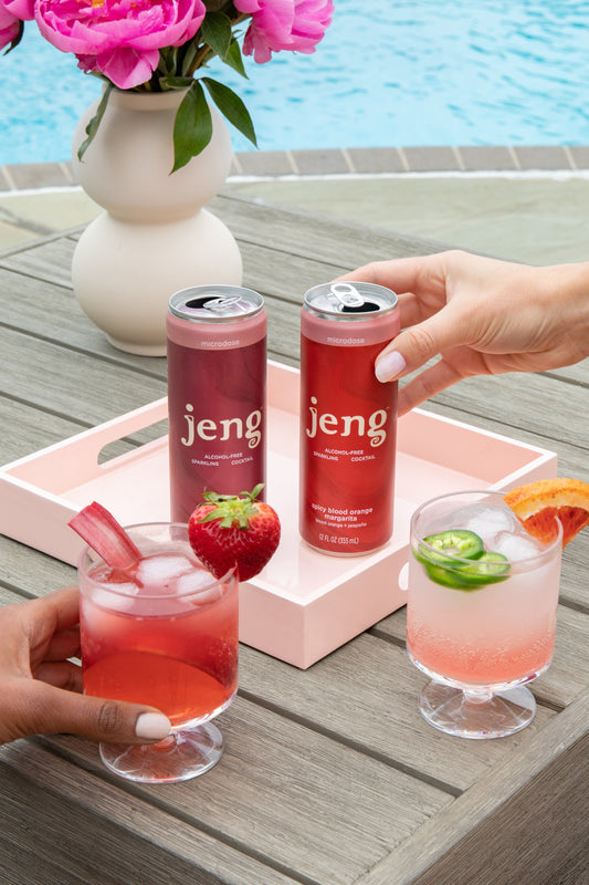 Two Jeng zero-proof cocktail cans on a pink tray beside iced cocktails garnished with fresh strawberry and jalapeño.