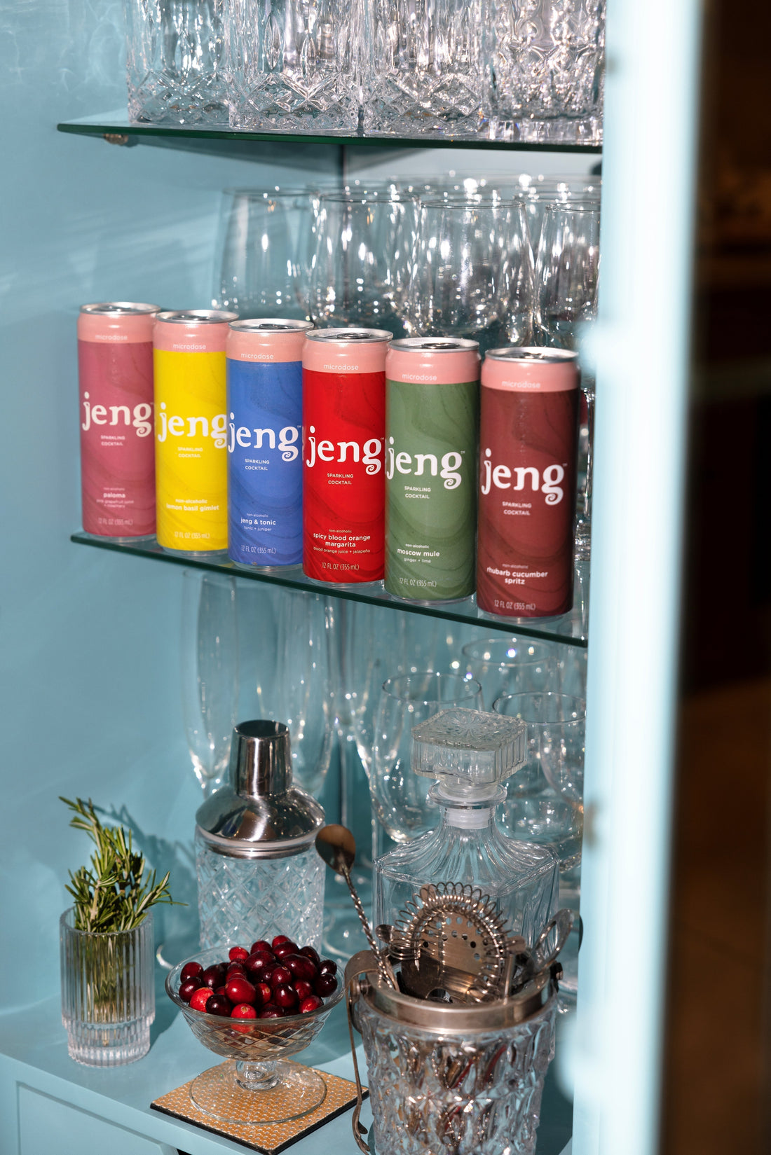 Six colorful cans of Jeng alcohol-free cocktails on a glass shelf with barware, cranberries, and a sprig of rosemary.