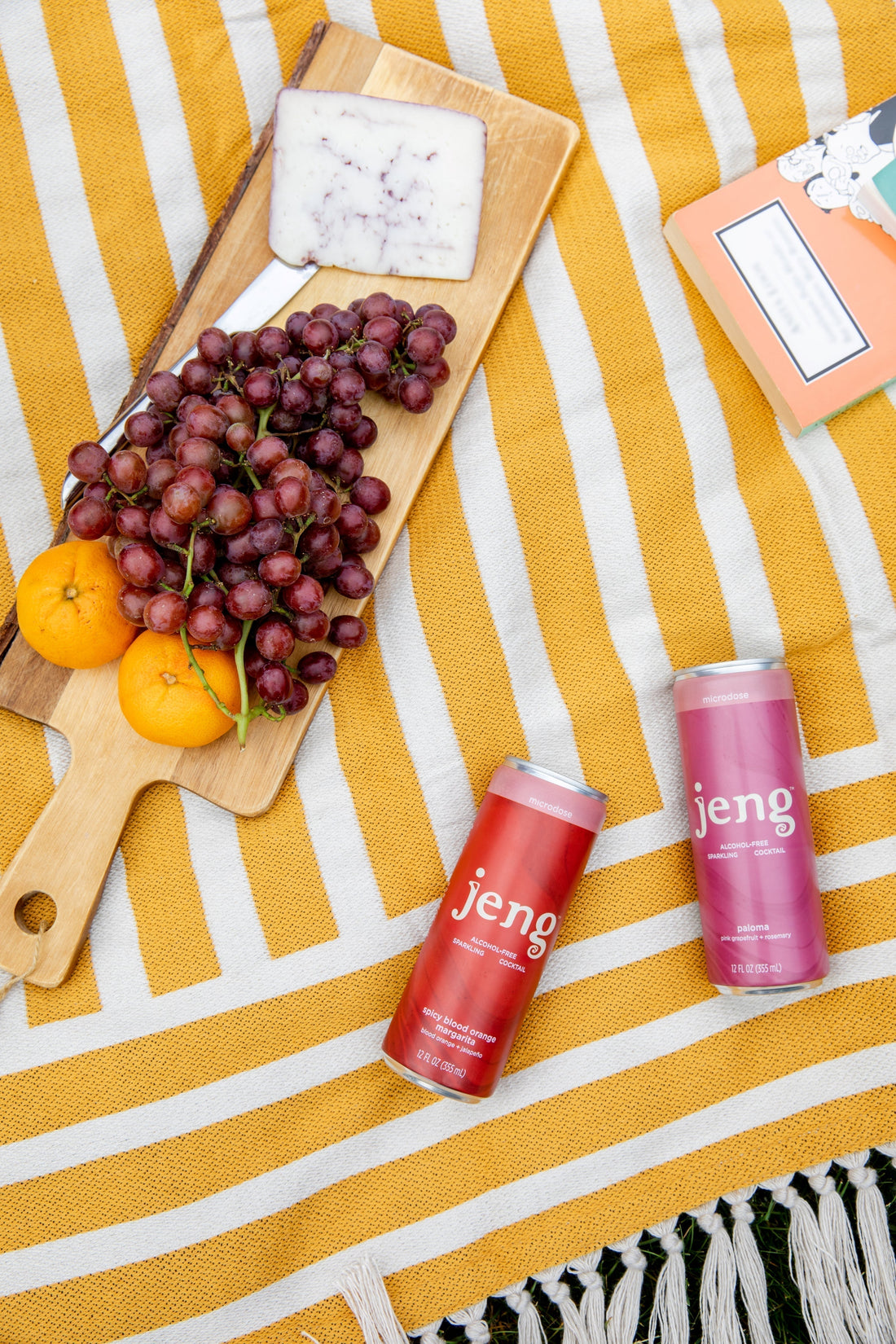 Jeng canned cocktails on a yellow striped blanket beside a charcuterie board with cheese, grapes, and oranges.