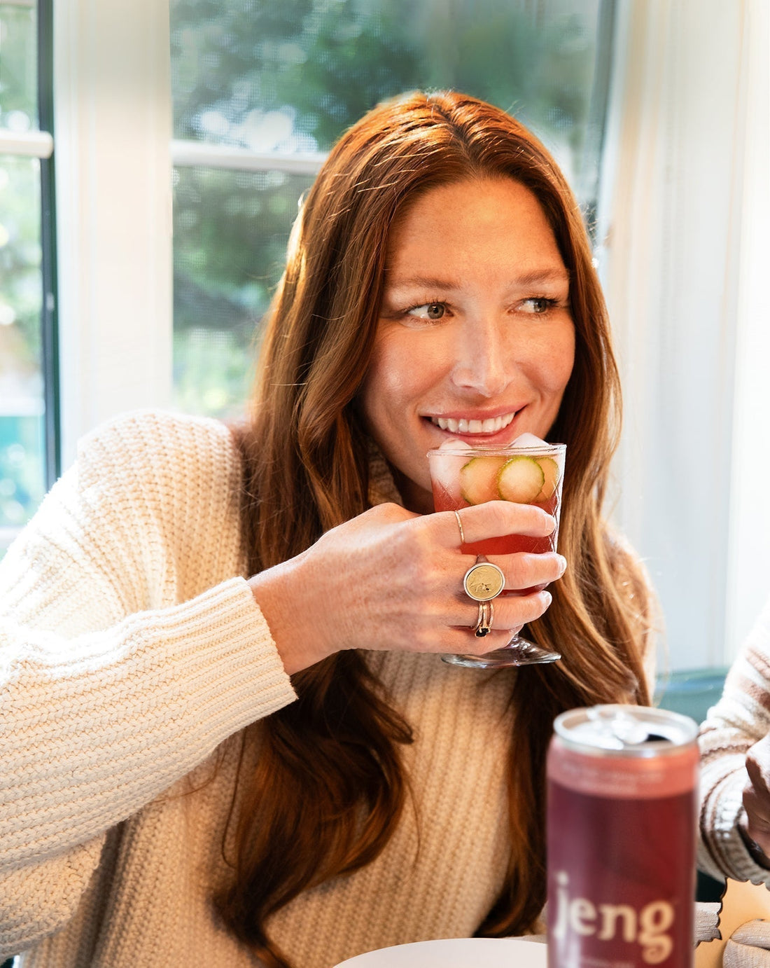 Smiling woman in a cozy sweater enjoying a chilled Jeng alcohol-free cocktail garnished with cucumber slices.