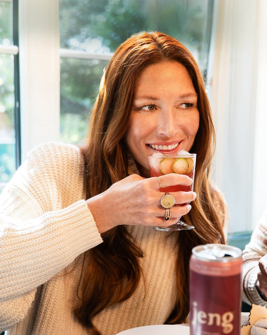 Smiling woman in a cozy sweater enjoying a chilled Jeng alcohol-free cocktail garnished with cucumber slices.