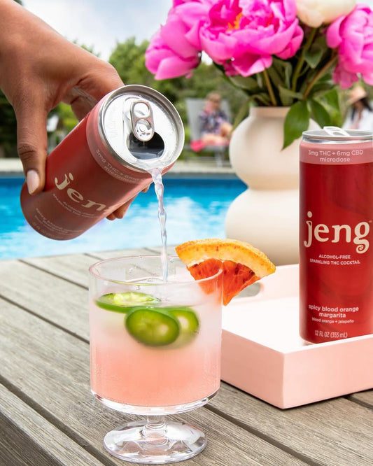 Hand pouring Jeng's alcohol-free THC cocktail into a glass with lime and grapefruit garnish by the pool with flowers.