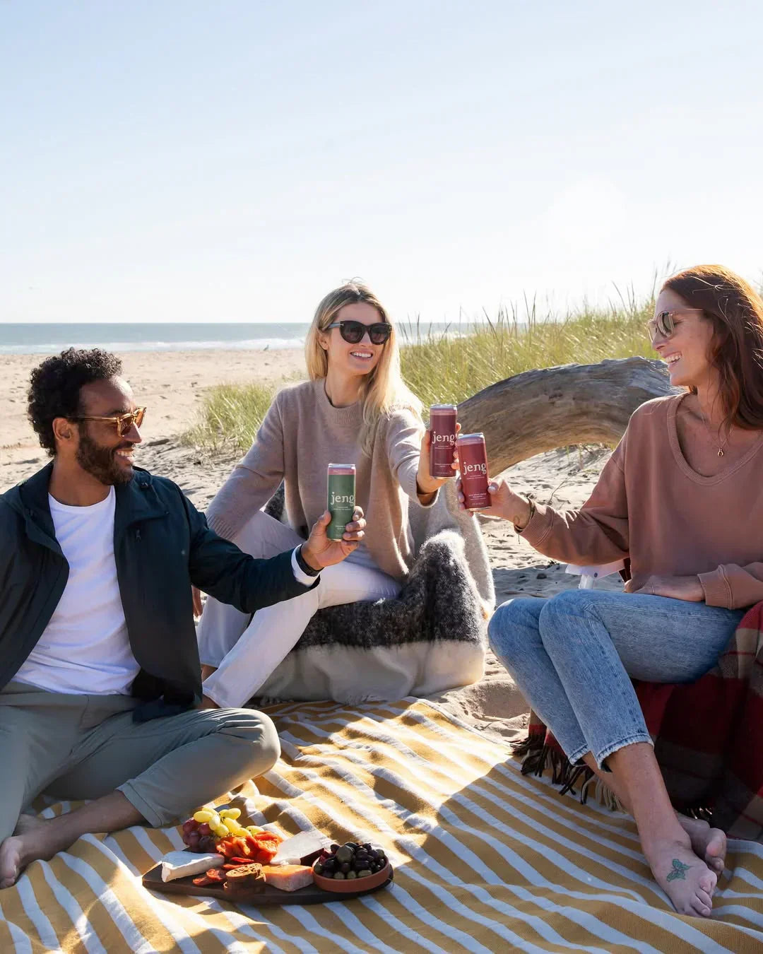 Three friends having a beach picnic and toasting with cans of Jeng cocktails.