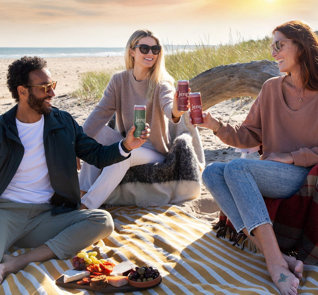 Three friends enjoying Jeng's alcohol-free cocktails on a beach blanket with a dog, sunset picnic setting.