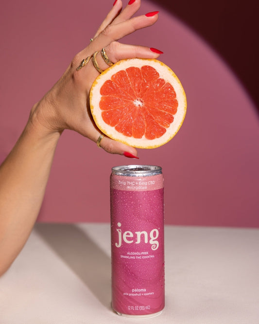 A hand holding a slice of grapefruit above a cold can of Jeng Paloma Sparkling THC Cocktail, covered in condensation.