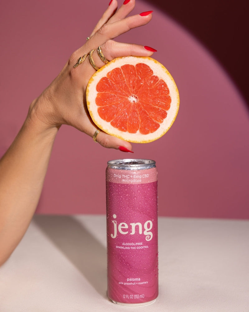 A hand holding a slice of grapefruit above a cold can of Jeng Paloma Sparkling THC Cocktail, covered in condensation.