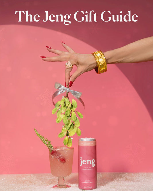 A hand holding mistletoe above a Jeng Paloma can and a garnished cocktail on a pink holiday-themed background.