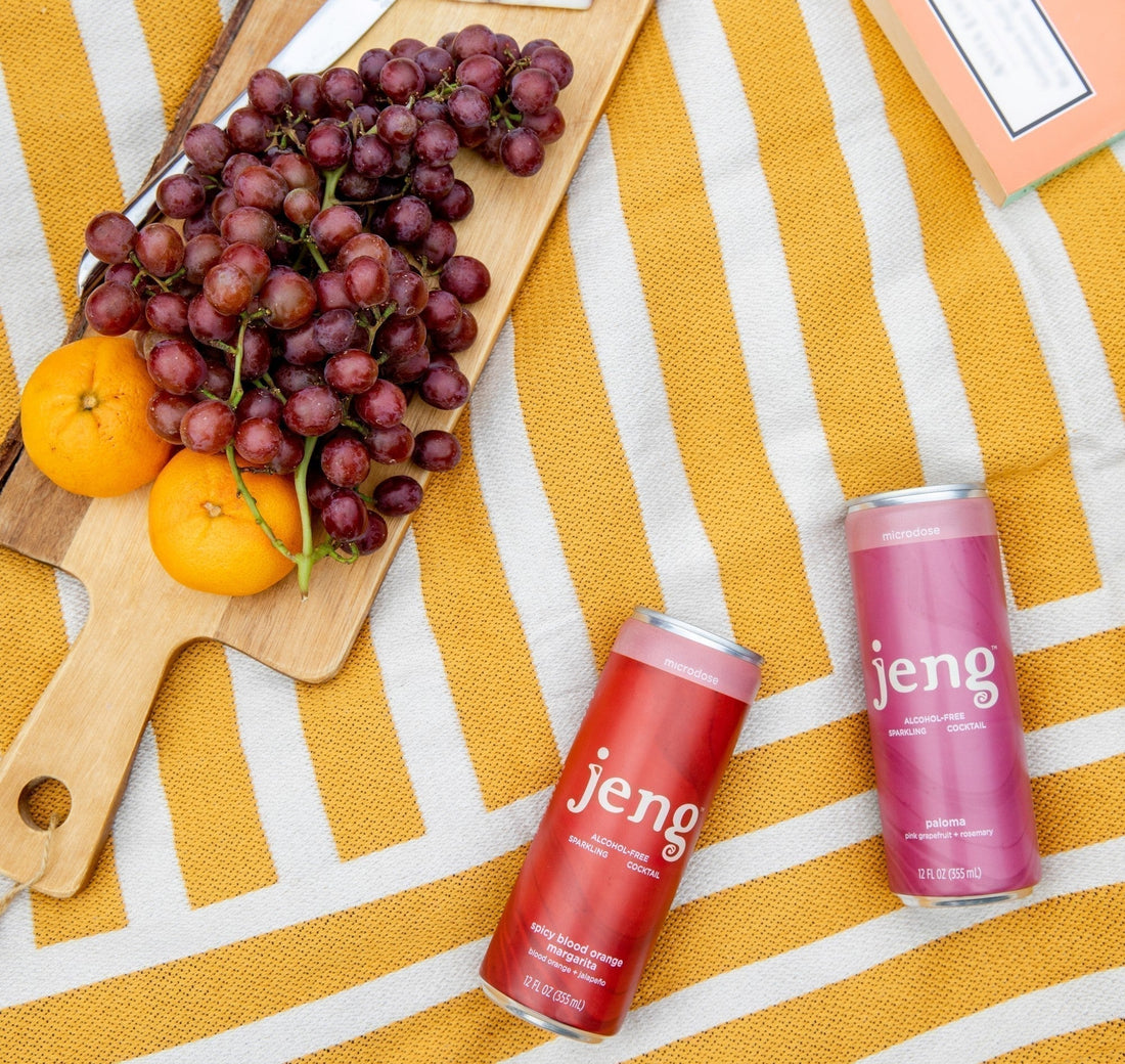 Jeng cans on a striped yellow picnic blanket with grapes, cheese, tangerines, and a book nearby.
