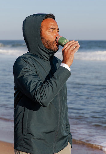 Man in a hooded jacket drinking a Jeng THC-infused beverage by the ocean shore.