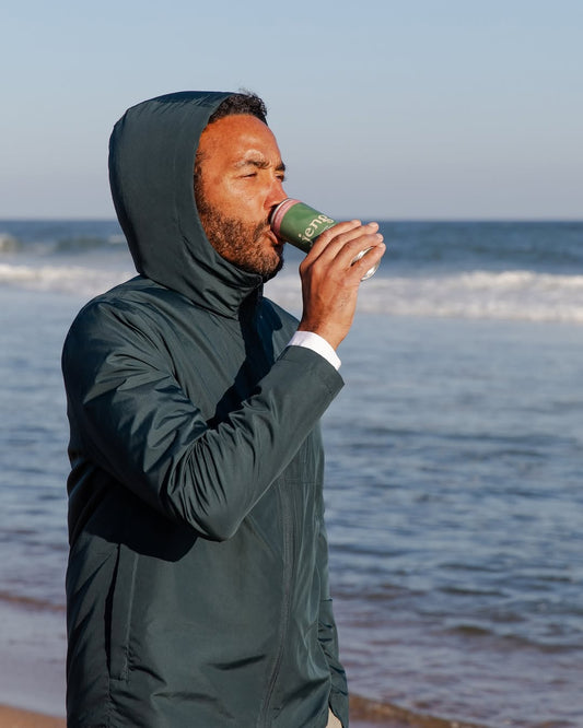 Man holding a sparkling non-alcoholic drink while standing by the ocean at sunset.