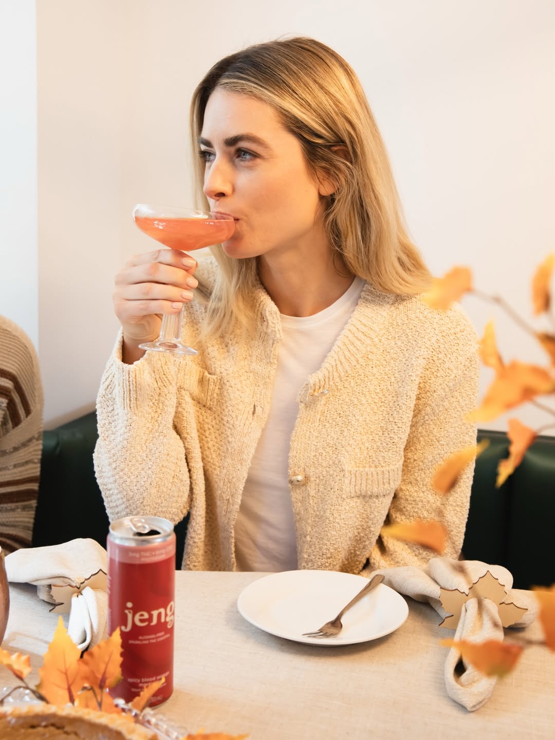Woman relaxing with a pink Jeng Spicy Blood Orange Margarita can beside a matching cocktail in a softly lit evening setting.