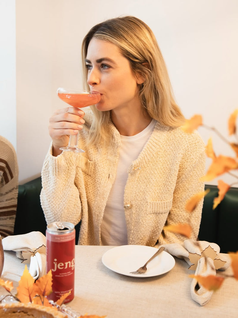 Woman relaxing with a pink Jeng Spicy Blood Orange Margarita can beside a matching cocktail in a softly lit evening setting.