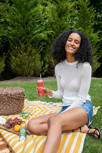A woman smiling and holding a Jene beverage can while sitting on a yellow-striped picnic blanket with grapes and fruits.